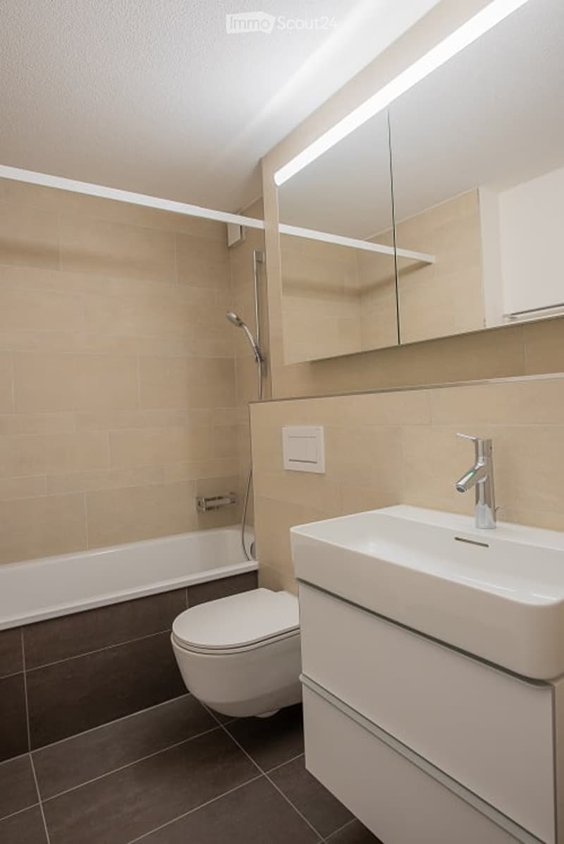 Modern bathroom with beige tiles, a bathtub, toilet, sink, and a large mirror with integrated lighting.