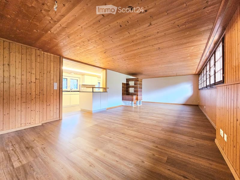 Empty room with wooden walls, ceiling, and floor, featuring large windows and an open doorway to another room with cabinets and a counter.