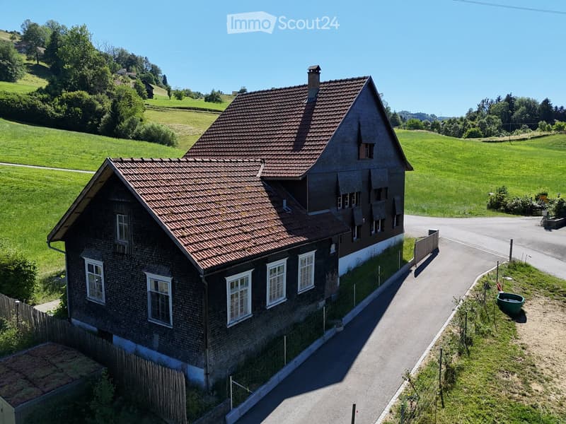 A two-story wooden house with a red-tiled roof, white-framed windows, and a small chimney, situated in a rural, green landscape with hills and trees in the background.