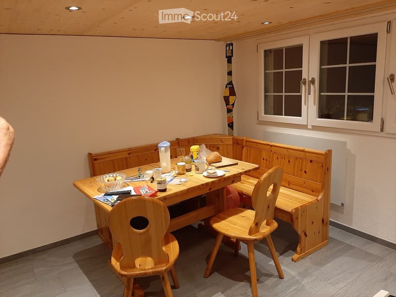 Cozy wooden dining area with a long bench seating, a wooden table, and two chairs. The table is set with cups, a bowl of fruit, and bread.