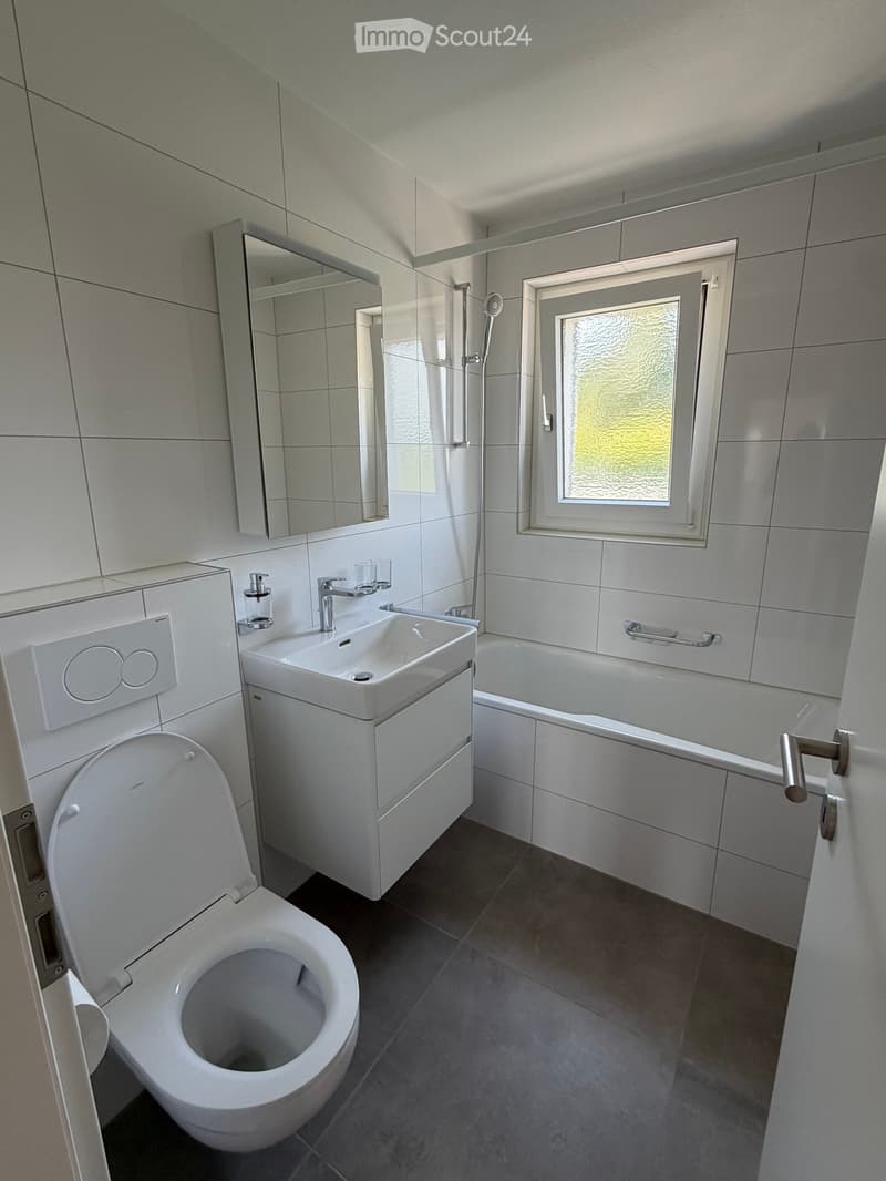 Modern bathroom with white tiles, a toilet, a sink with a mirror, a bathtub, and a shower area. The room features a frosted window and a sleek, contemporary design.