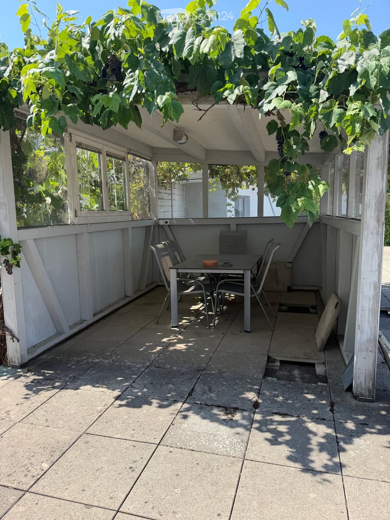 A covered patio with a table and chairs, surrounded by grapevines and white wooden railings.