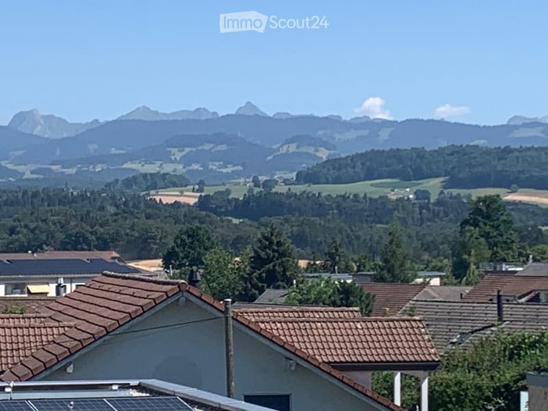 A panoramic view of a residential area with tiled roofs, surrounded by dense forests and distant mountains under a clear blue sky.