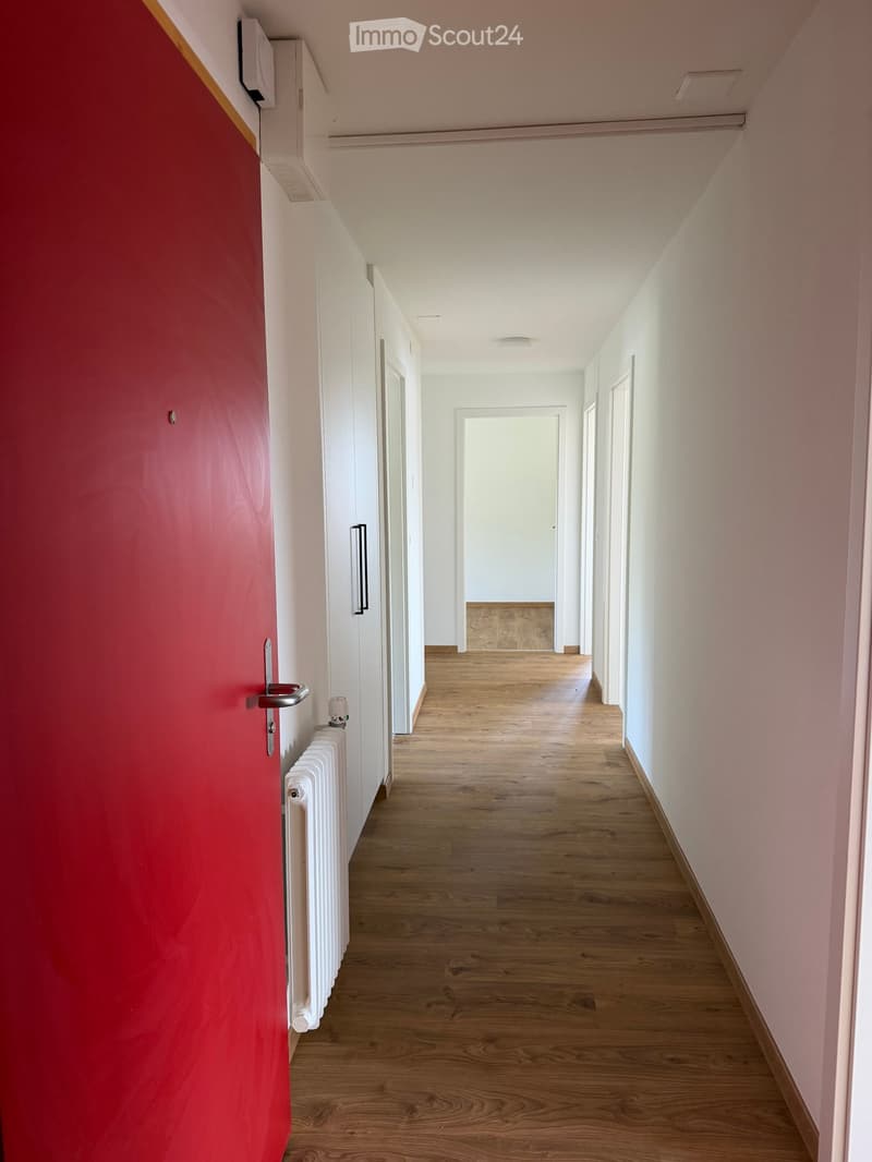 Empty hallway with wooden flooring, white walls, and a red door on the left side.