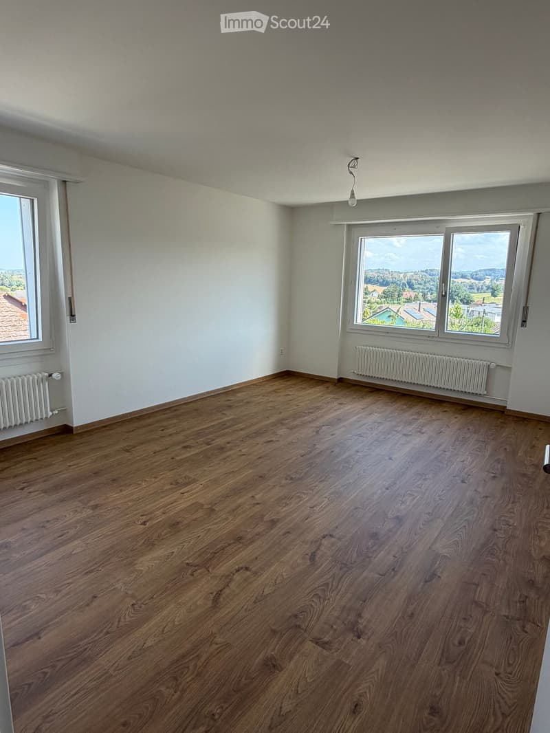 Empty room with wooden flooring, white walls, and large windows offering a scenic view of the countryside.