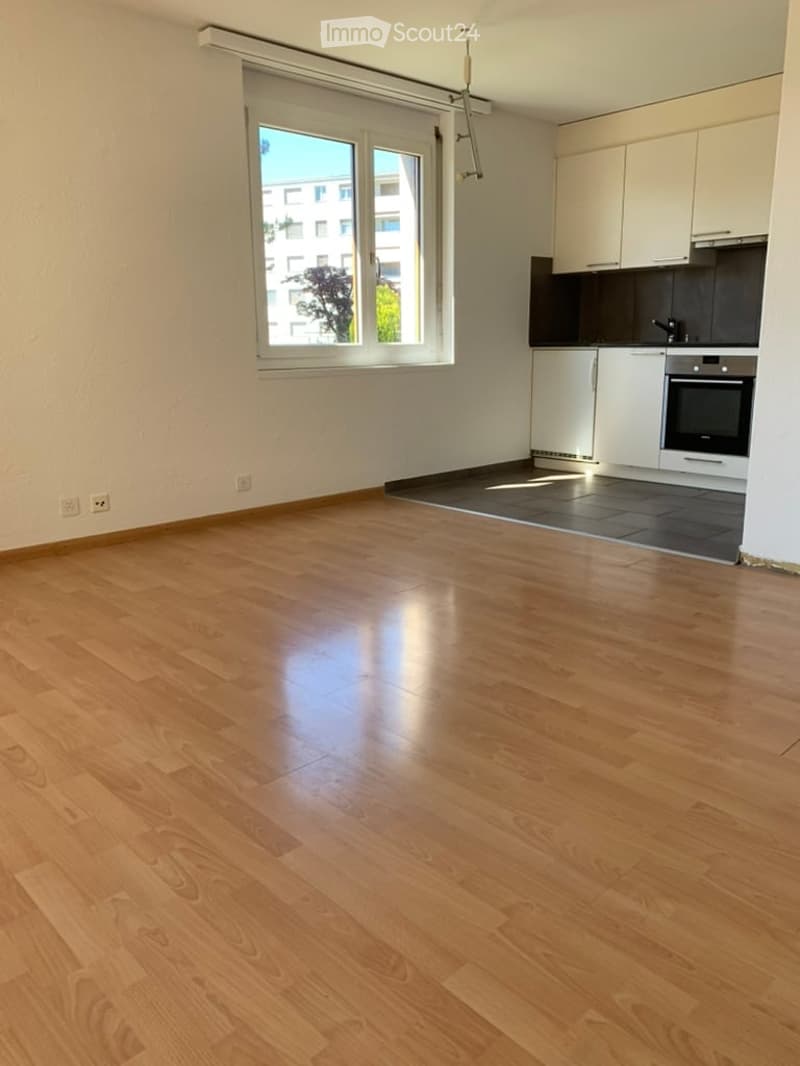 Empty room with wooden flooring, white walls, and a small kitchen area with cabinets and appliances visible in the background.