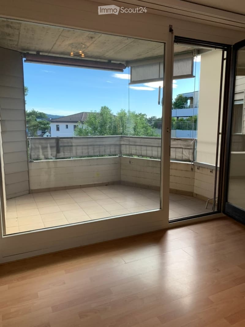 Empty room with wooden flooring and large sliding glass doors opening to a tiled balcony area with a view of trees and a building.