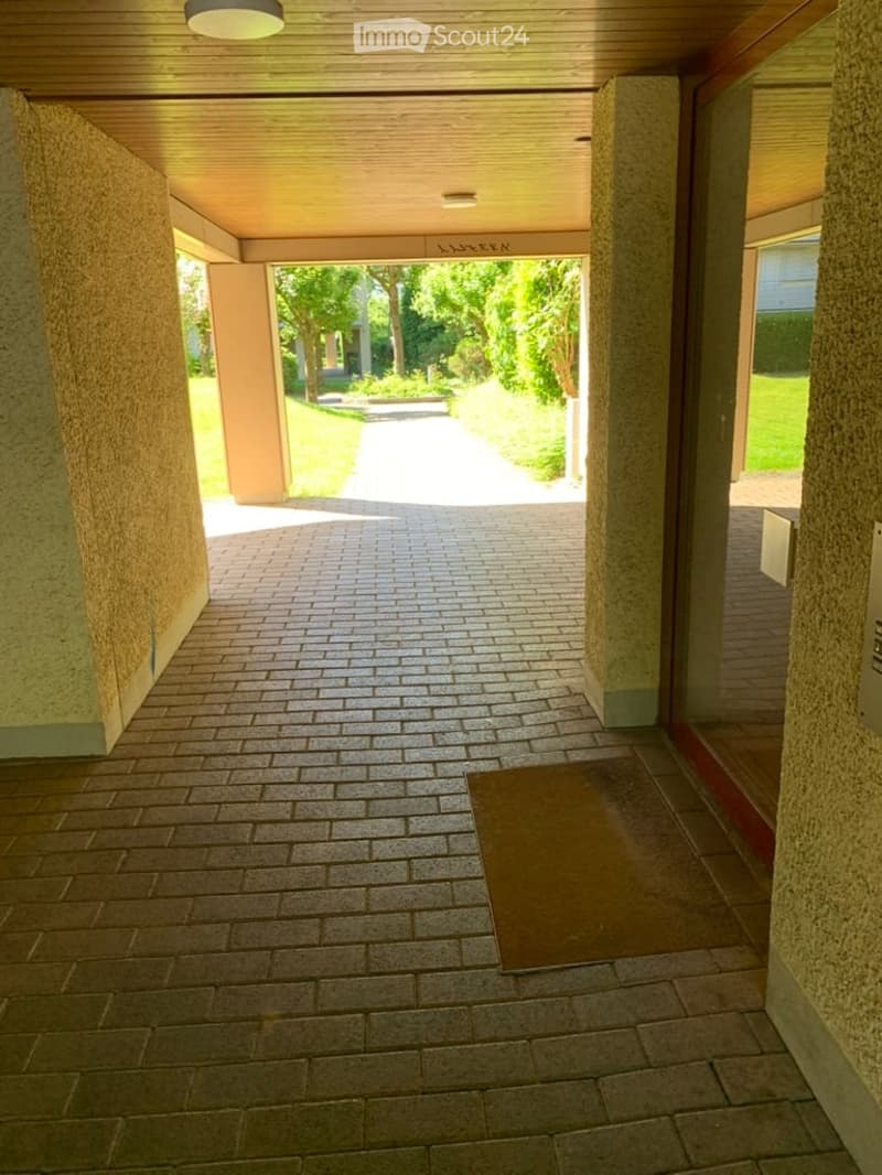 Brick-paved hallway with wooden ceiling and recessed lighting, leading to an open area with greenery visible outside.