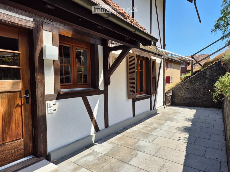Traditional wooden-framed building with white plaster walls, wooden shutters, and a tiled roof, situated on a raised ground floor with a paved pathway.