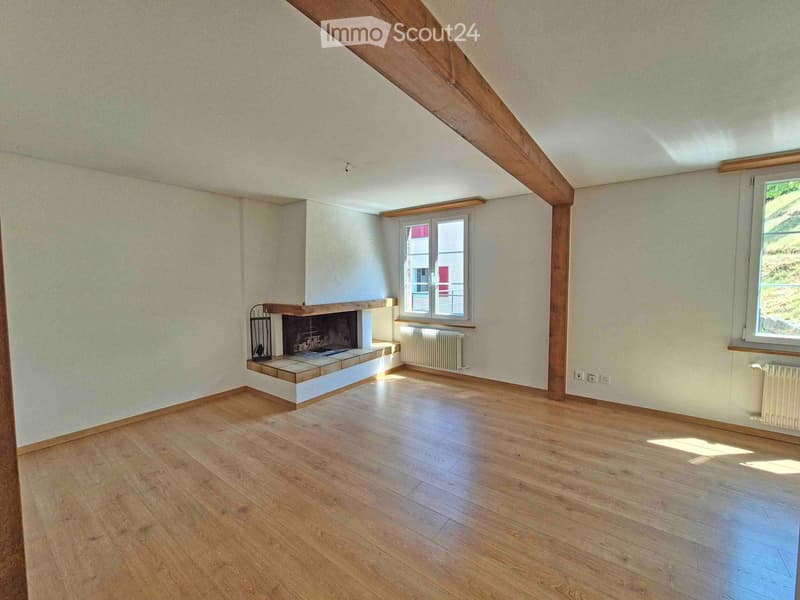 Empty room with wooden flooring, exposed wooden beams, fireplace, and two windows letting in natural light.
