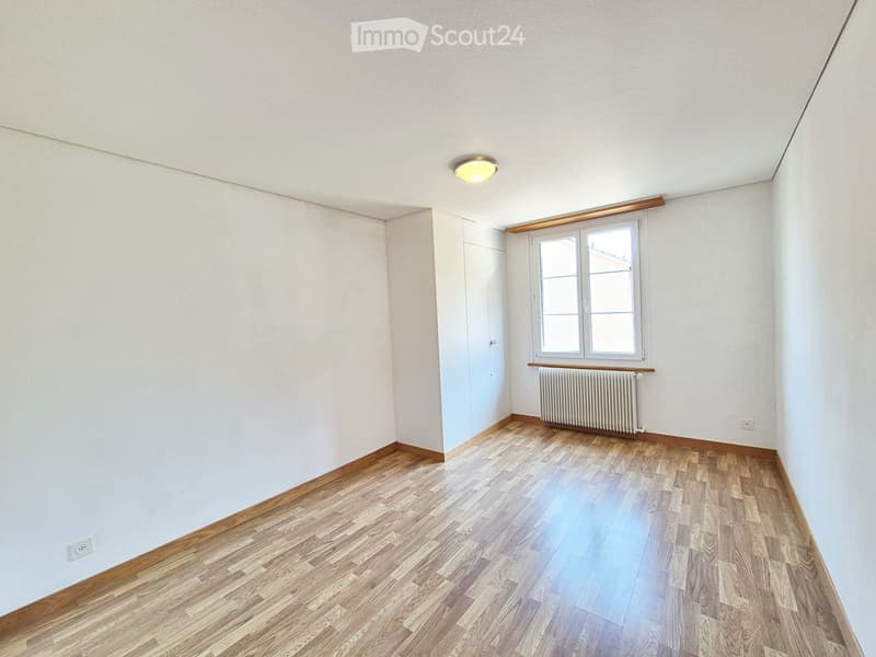 Empty room with light-colored walls, wooden flooring, a window with white frames, a radiator, and a ceiling light fixture.