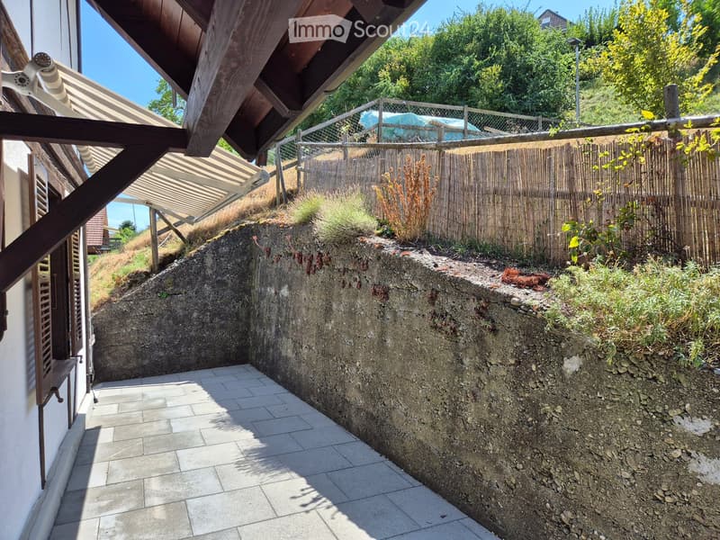 A raised garden area with a stone retaining wall, small plants, and a wooden fence. The area is paved with tiles and has a wooden pergola structure on the left side.