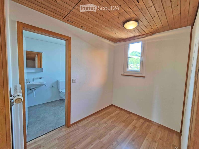 Bathroom with wooden flooring, white walls, a window, a sink, a toilet, a mirror, and a fan. The adjacent room is also visible.