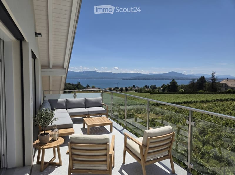 Balcony with a modern glass railing, overlooking a scenic lake and mountains. Features a grey sectional sofa, wooden coffee table, two wooden chairs, and a small potted plant.