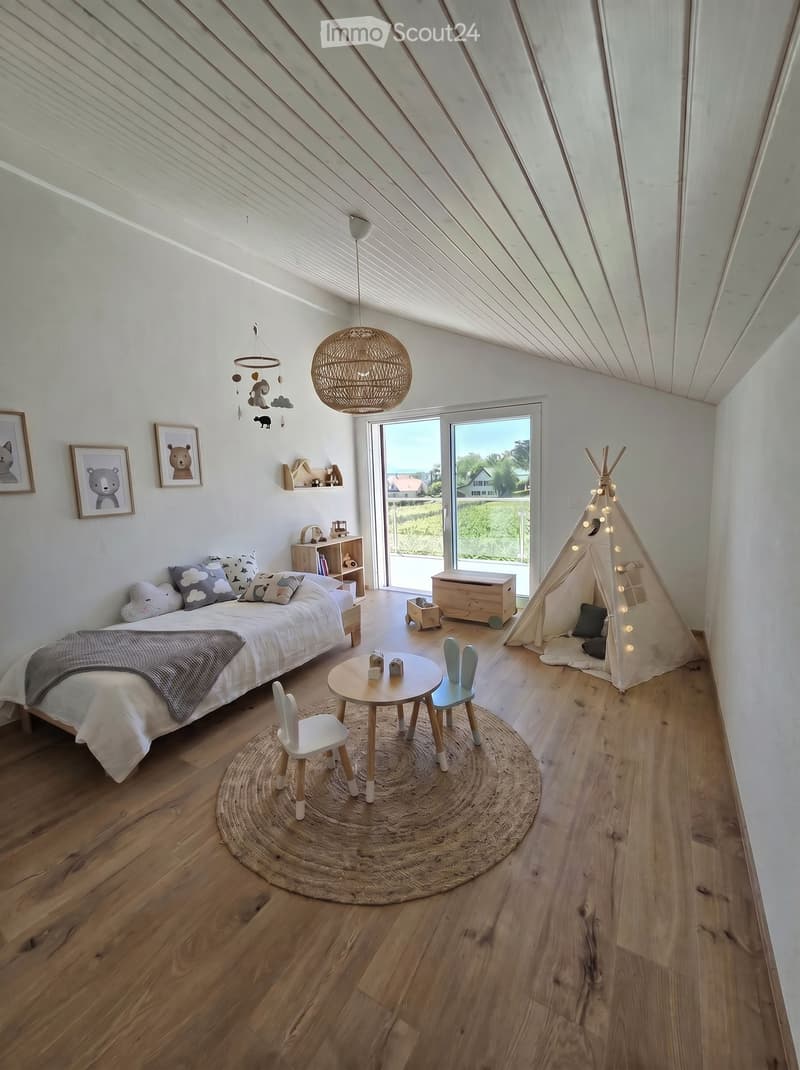 Bright, airy bedroom with wooden floors, a large bed, a small round table with chairs, a teepee tent, and a hanging wicker lamp. Large windows offer a view of the outdoors.