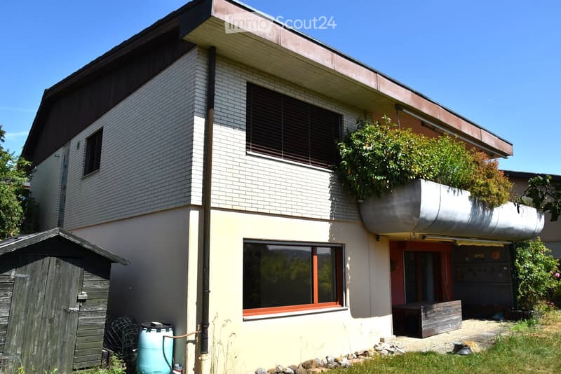 Two-story house with white brick facade, large windows, and a balcony with plants. Features a wooden shed and a water tank in the yard.