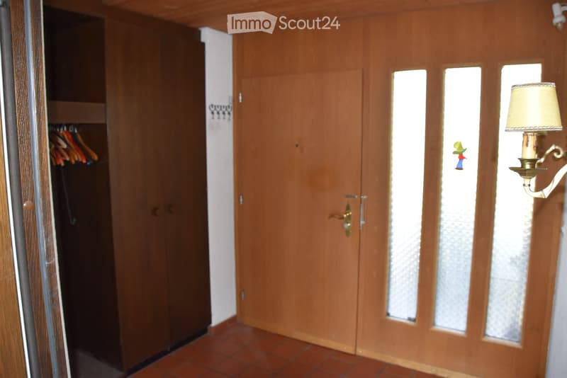 A narrow hallway with wooden doors, a closet with hanging clothes, and a wall-mounted lamp. The floor is tiled, and the walls are painted white.