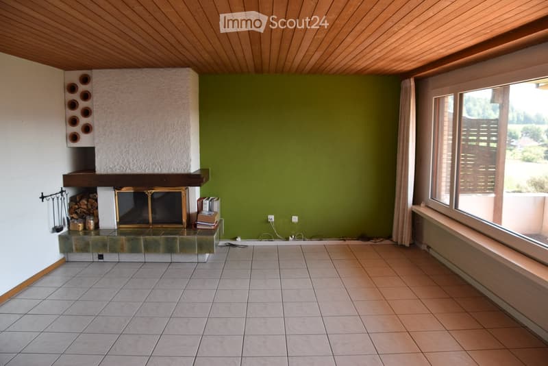 Empty room with tiled floor, green wall, large window, and fireplace with wood stack and tools.