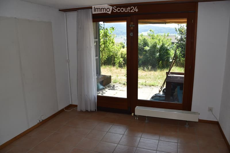 Empty room with tiled floor, large window with wooden frame, white curtain, radiator, and view of outdoor area with greenery.