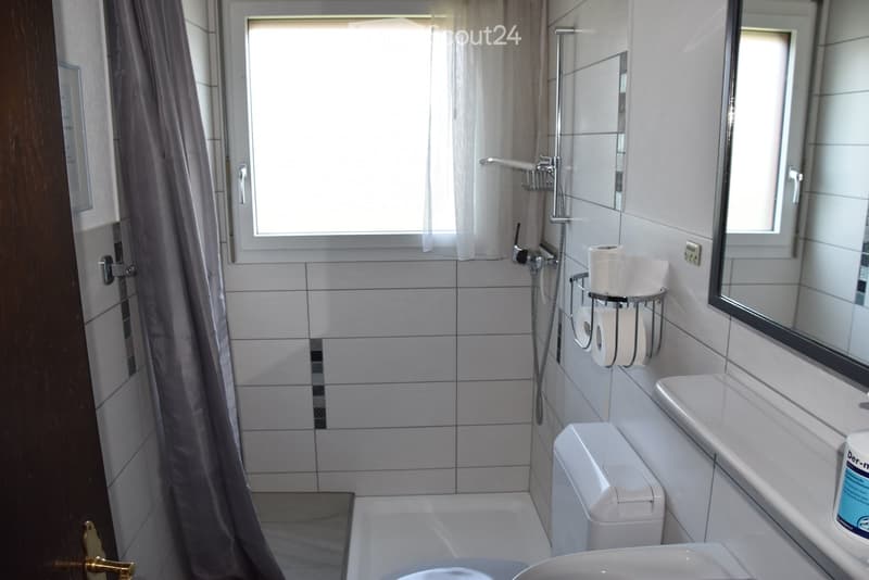Modern bathroom with white tiles, a bathtub, toilet, sink, mirror, and a shower with a curtain. Toilet paper is stored on a wall-mounted holder.