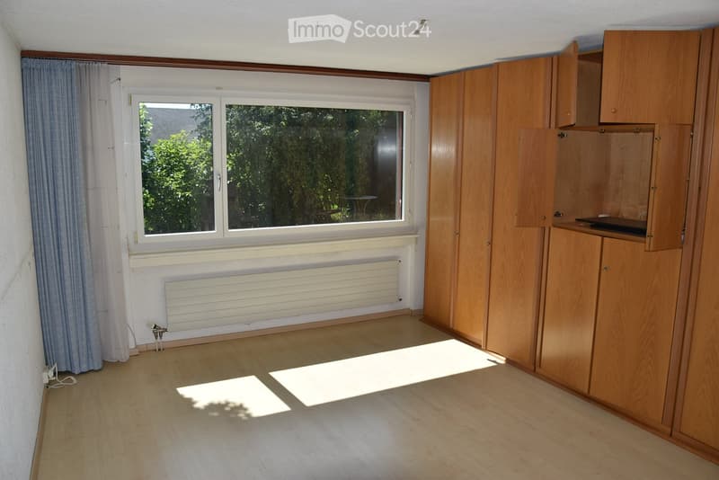 Empty room with wooden flooring, white walls, large window with curtains, built-in wooden cabinetry, and radiator under the window.