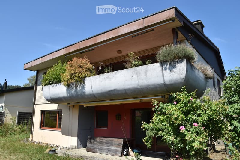 Two-story house with a modern design, featuring a large balcony with plants, a red front door, and a wooden bench near the entrance.