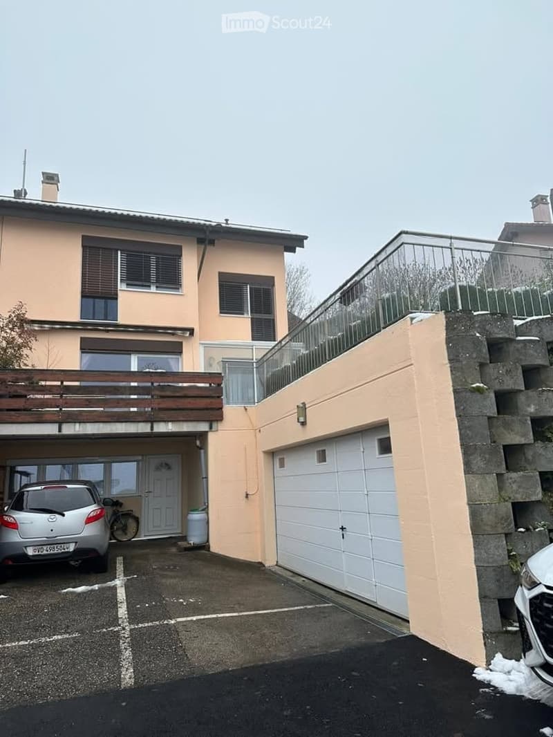 Two-story house, beige exterior, balcony with wooden railing, garage, car parked in the driveway