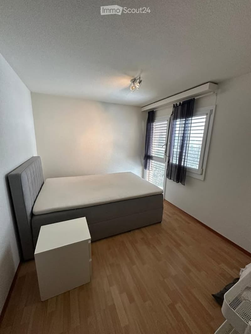 Single bed, wooden floor, white walls, ceiling lights, nightstand, window with blinds, white storage unit
