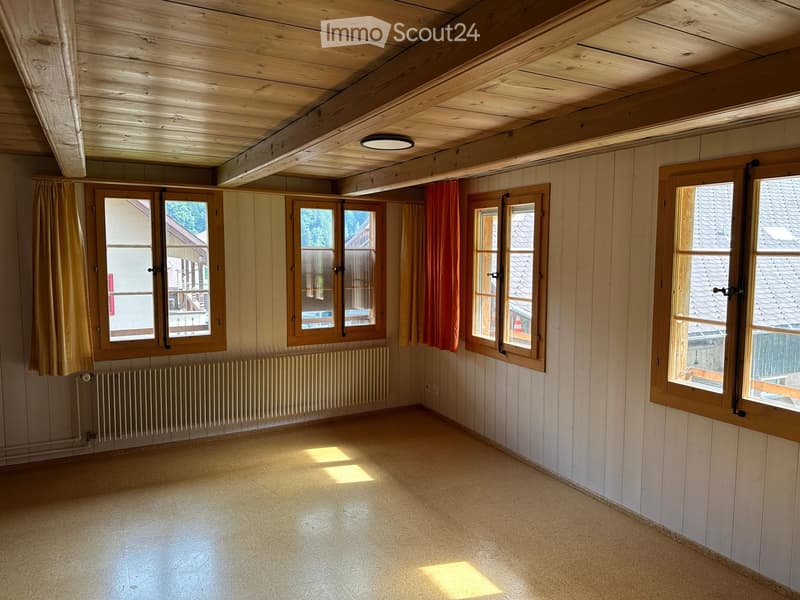 Empty room with wooden walls, exposed wooden ceiling beams, large windows with curtains, and a radiator. No furniture or equipment present.