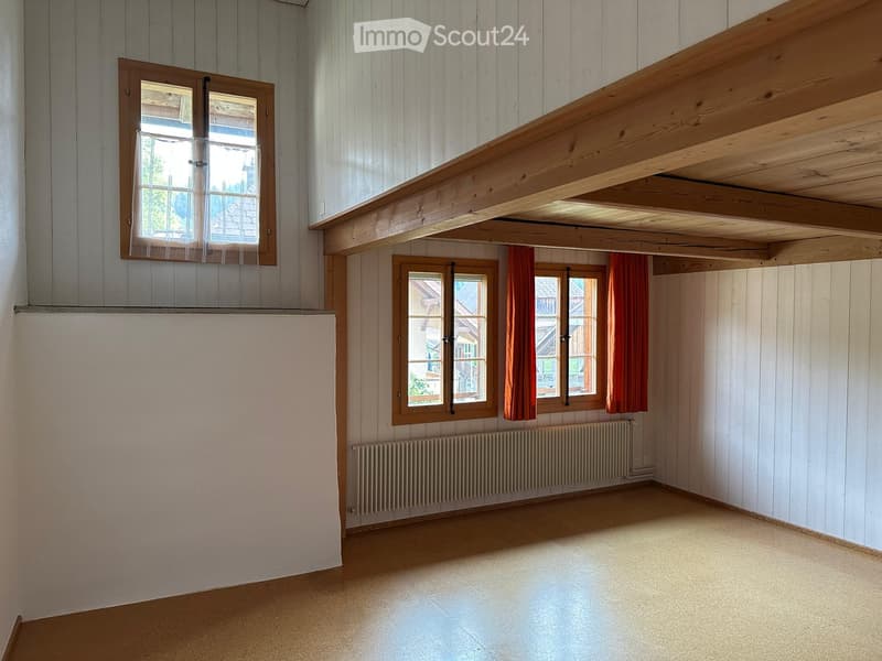 Empty room with wooden walls, exposed wooden beams, and large windows with red curtains. The floor is light-colored and the room has a radiator under the windows.