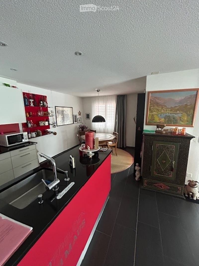 Modern kitchen with red and black cabinets, a black countertop, a sink with a modern faucet, a microwave, and a dining area with a table and chairs. A colorful picture frame and a small dog are also visible.