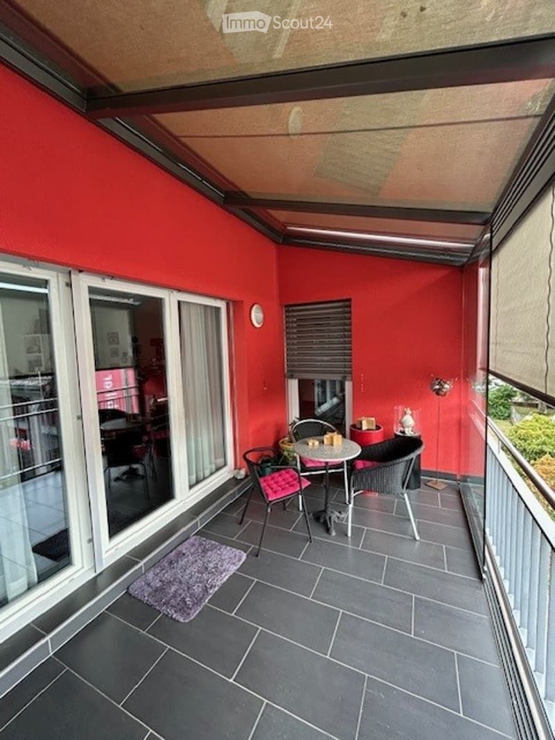 Balcony with red walls, grey tiled floor, small round table with chairs, purple rug, and a view of the interior space through large glass doors.