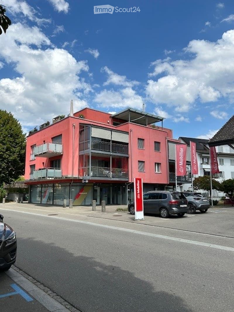 Modern red building with multiple floors, balconies, and a storefront on the ground level. Several cars are parked in front.