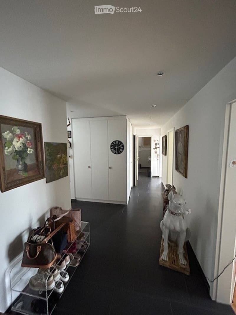 A long, narrow hallway with white walls, black tiled flooring, and several framed artworks. There is a white cabinet at the end of the hallway, and a small shoe rack with shoes and bags on the left side.