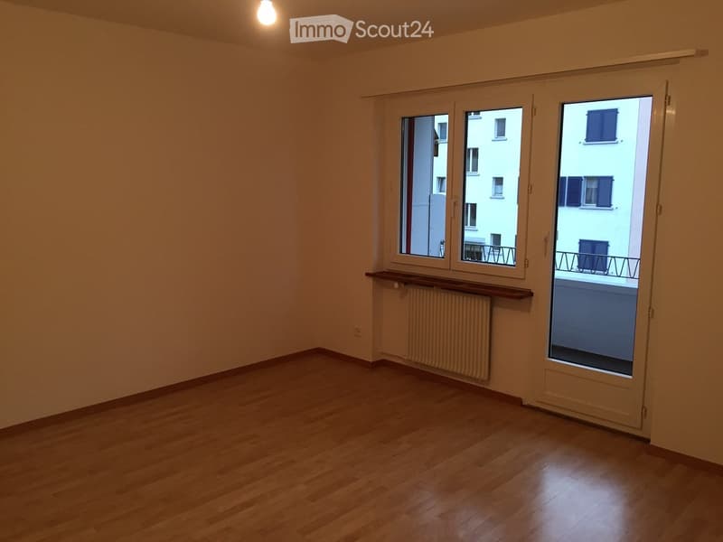 Empty room with wooden flooring, large windows, and a radiator. The room has a balcony accessible through a sliding door.