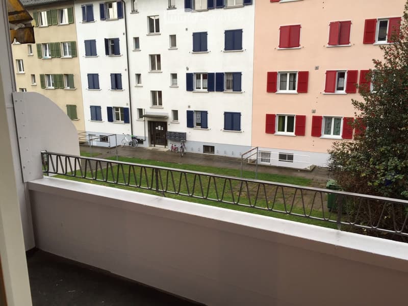Balcony overlooking a small courtyard with green grass, metal railing, and several residential buildings with colorful shutters in the background.