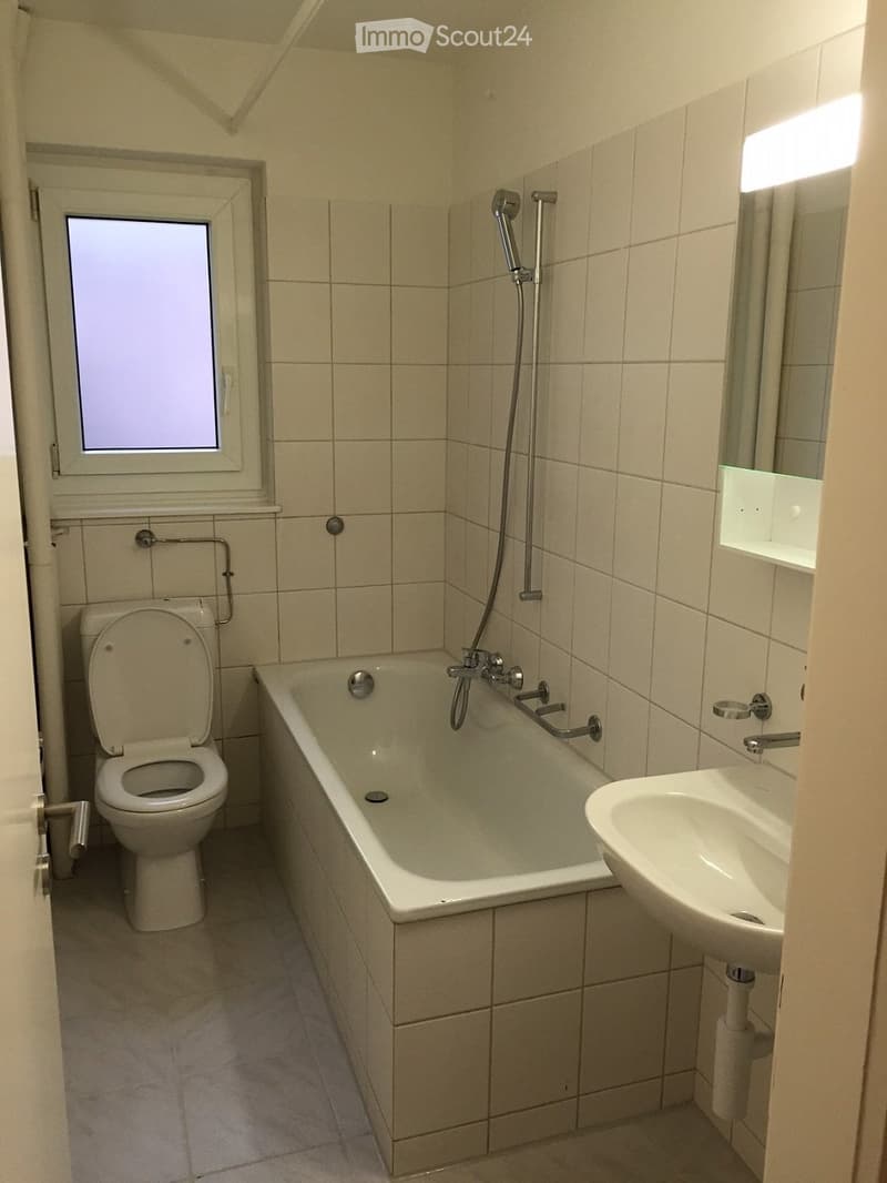 Bathroom with a toilet, bathtub, sink, and mirror. The walls are tiled, and there is a window with frosted glass.