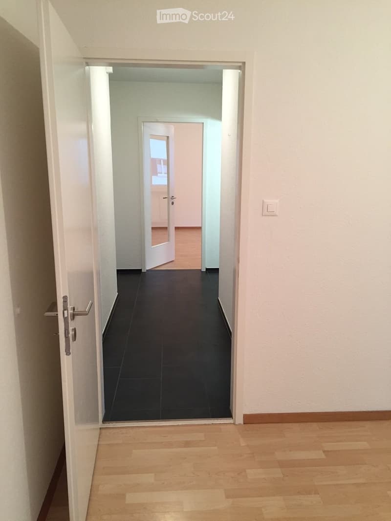 Empty hallway with wooden flooring, white walls, and a black tiled floor in the adjacent room. The hallway features a white door with a silver handle and a visible light switch on the right wall.