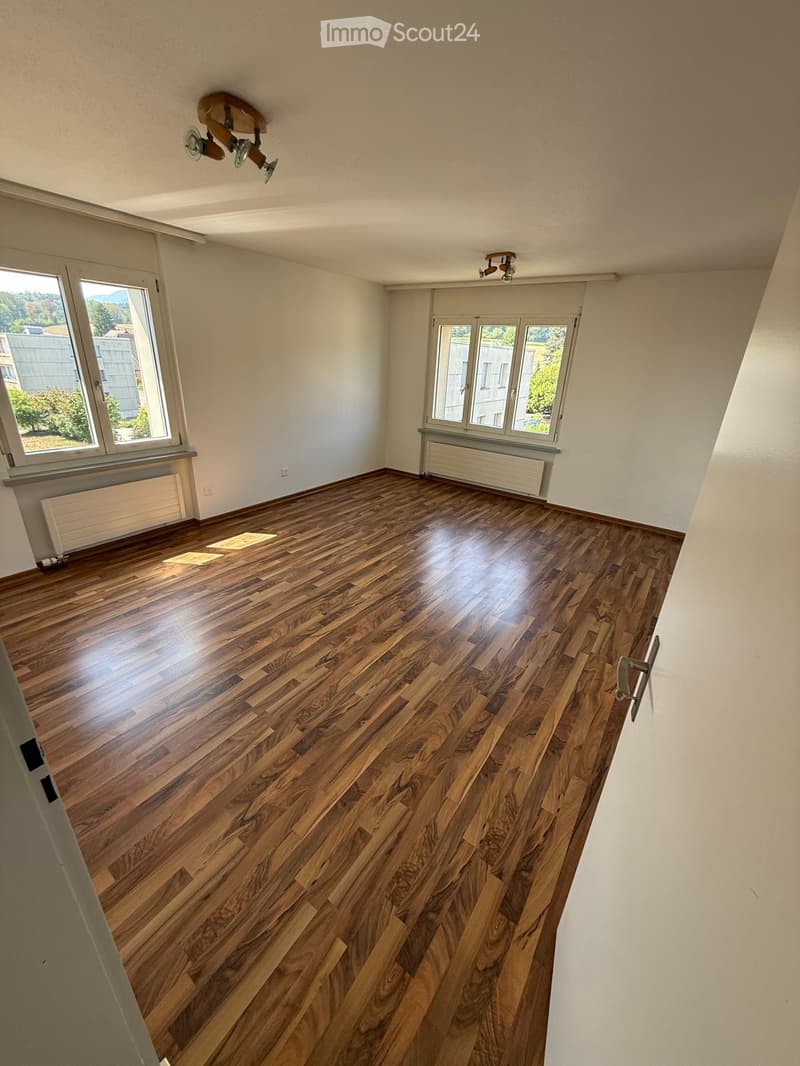 Empty room with wooden flooring, white walls, large windows, and ceiling lights.