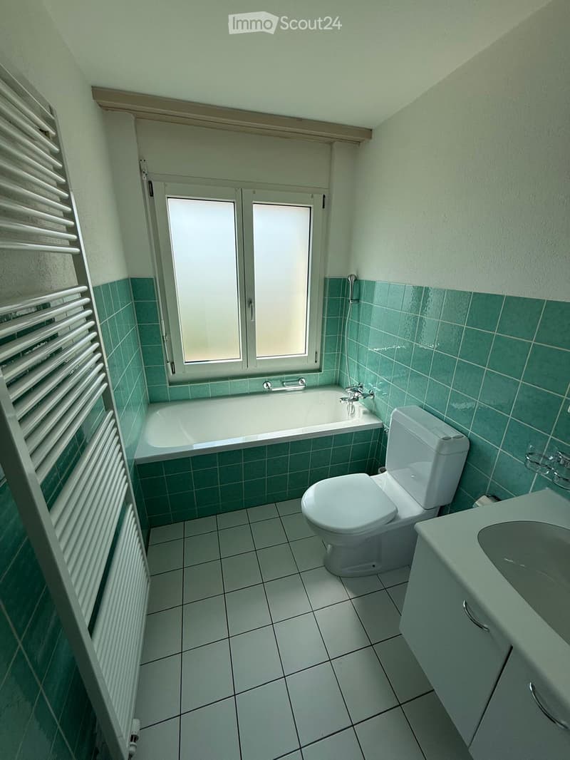 Bathroom with green tiled walls, white bathtub, toilet, sink, and radiator. Features a window with frosted glass panes.