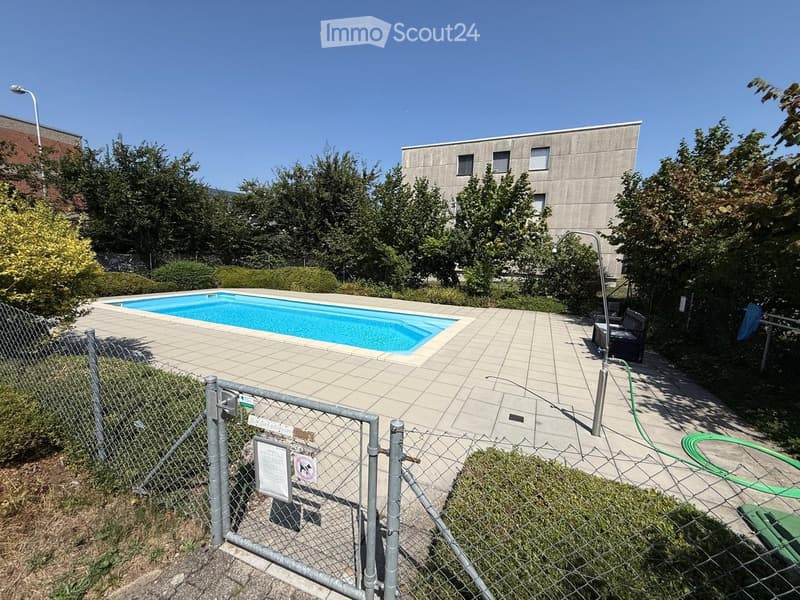 Outdoor area with a swimming pool, tiled deck, surrounding greenery, and a modern building in the background.