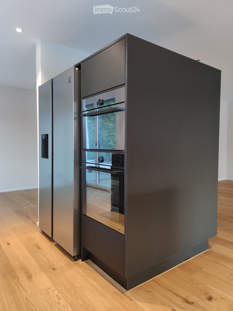 Modern kitchen with stainless steel refrigerator and built-in oven, light wooden flooring, and white walls.