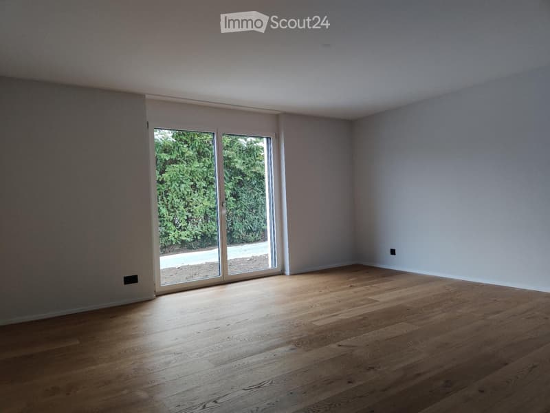 Empty room with wooden flooring, large sliding glass doors leading to a green outdoor area, and white walls.