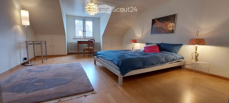 Bright bedroom with wooden floors, slanted ceiling, large window, bed with blue bedding, side table with lamp, chair, small table, and artwork on the wall.