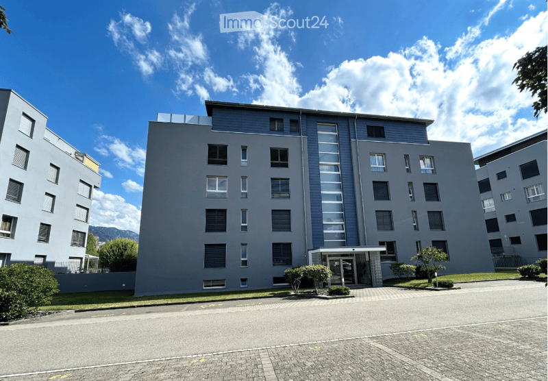 Modern multi-story apartment building with grey facade, large windows, and a small landscaped entrance area.