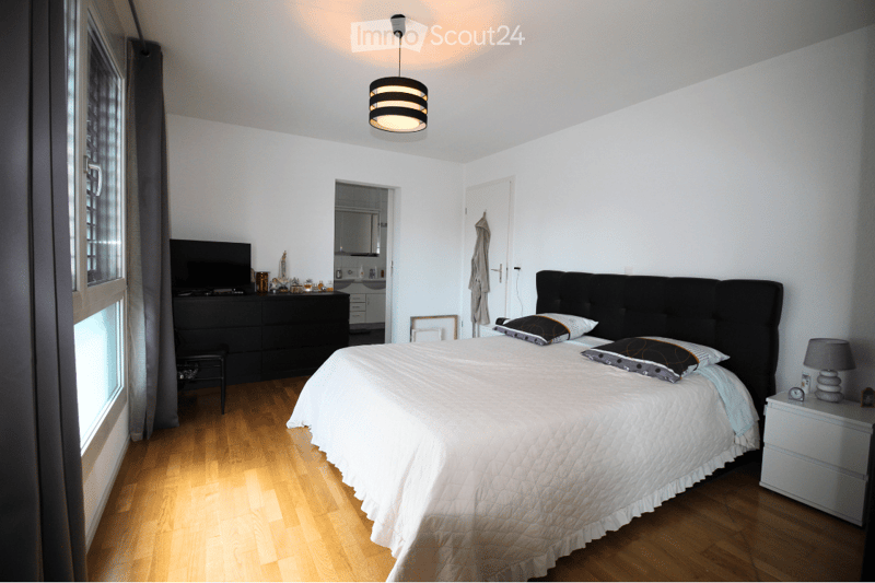 Bright bedroom with wooden flooring, large bed with white bedding and decorative pillows, nightstand with lamp, dresser, chair, and a small table with items.