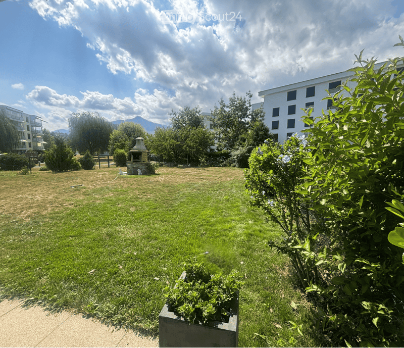 A spacious, well-maintained garden area with green grass, trees, shrubs, and a small stone fireplace in the background. The area is bordered by a modern building and features a clear, partly cloudy sky.