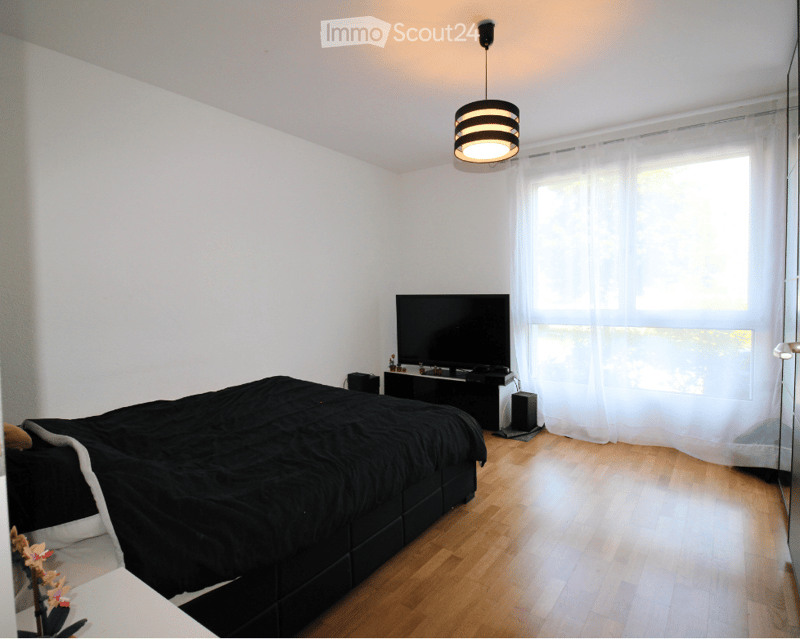 Modern bedroom with wooden flooring, large window with white curtains, bed with black bedding, television on a stand, and a hanging lamp.