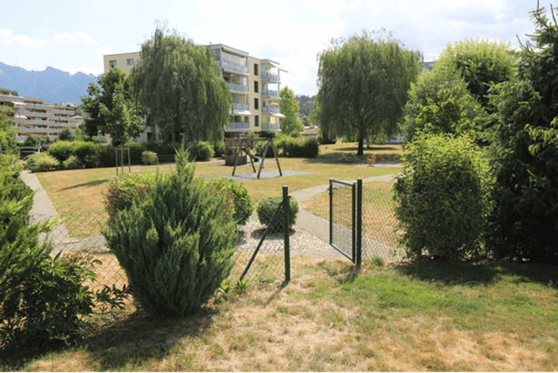 Well-maintained garden area with trimmed bushes, trees, a small playground structure, and a pathway leading to a gated entrance.