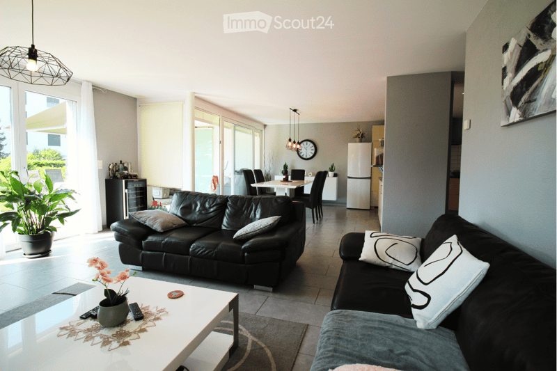 Modern living room with black leather sofas, a white coffee table, a dining area with chairs and a table, a refrigerator, and a potted plant near the window.